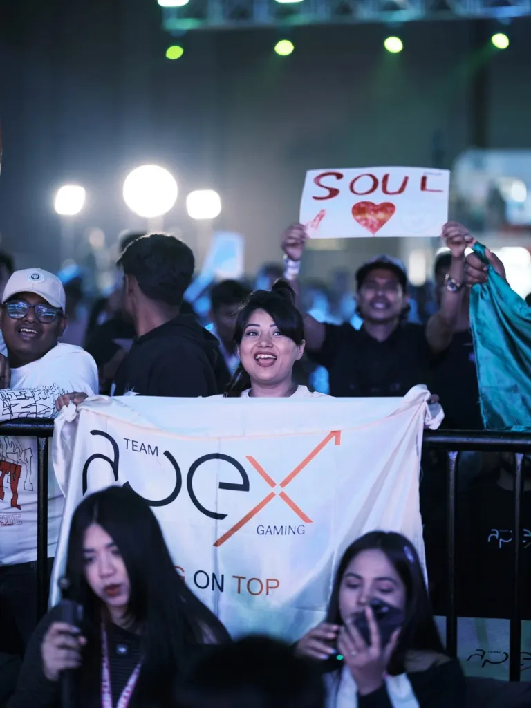 A Team Apex Gaming supporter cheers from the barricade while a Team Soul fan holds a sign high behind her. (Image via KRAFTON India)